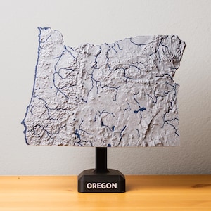 Puede incluir: Un mapa gris tridimensional de Oregón, con líneas azul oscuro que representan ríos y lagos. El mapa está montado sobre un soporte negro con el nombre del estado en letras blancas. Se exhibe sobre una superficie de madera.