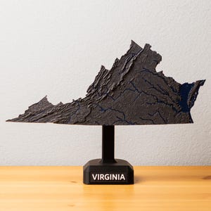 May include: A dark gray, textured sculpture of the state of Virginia, mounted on a black stand. The stand has the word "VIRGINIA" in white text. The sculpture has a detailed, topographical appearance.