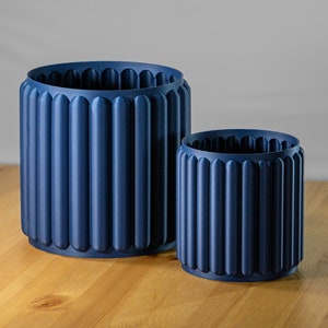 May include: Two blue ceramic planters with a ribbed design. The larger planter is in the background and the smaller planter is in the foreground.