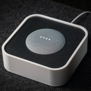 Może przedstawiać: Biały, kwadratowy dok do głośnika z szarym głośnikiem Google Home Mini w środku. Głośnik ma cztery białe lampki z przodu.