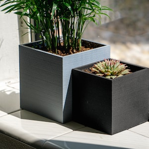 Könnte beinhalten: Zwei quadratische Blumentöpfe, einer grau und einer schwarz, stehen auf einer weißen, gefliesten Fensterbank. Eine grüne Pflanze mit langen Blättern befindet sich im grauen Topf, und eine Sukkulente im schwarzen Topf.