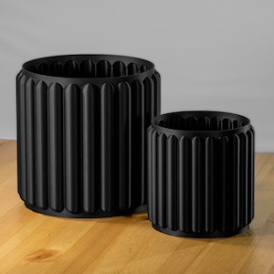 Pode incluir: Dois vasos cilíndricos pretos com um design nervurado. O vaso maior está no fundo e o menor está em primeiro plano.