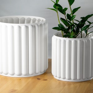 May include: Two white, cylindrical planters with a fluted design. One is empty, the other contains a green plant. The planters are made of a solid, opaque material and have a clean, modern aesthetic.