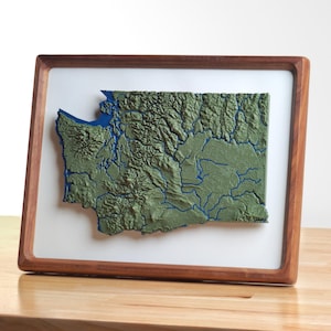 Può includere: Una mappa topografica 3D incorniciata dello stato di Washington caratterizzata da un paesaggio verde testurizzato con dettagli blu di fiumi e specchi d'acqua, su sfondo bianco all'interno di una cornice in legno.