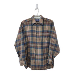 Peut inclure: Une chemise à carreaux à manches longues dans les tons de bleu, marron et bordeaux. La chemise a une fermeture boutonnée, une poche poitrine et des poignets boutonnés. La chemise est suspendue à un cintre blanc.