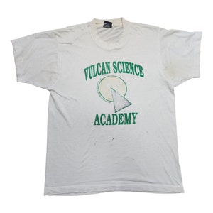 May include: White t-shirt featuring the green text "VULCAN SCIENCE ACADEMY". The design includes a circle and a triangle. The shirt has a crew neck and short sleeves. The shirt shows signs of wear.