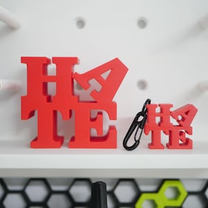 May include: Two red 3D printed words, "Hate" and "Hate", are displayed on a white surface. The larger word is made of individual letters, while the smaller word is a single piece. A black keychain is attached to the smaller word.