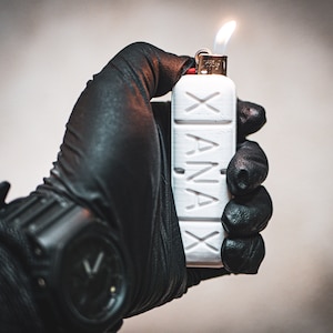 以下が含まれることがあります： 「XANAX」の文字がプリントされた白いライターが、手袋をした手によって持たれています。ライターは点火されており、上部に炎が見えます。背景はぼやけており、ニュートラルな色調です。
