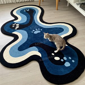 May include: A unique, abstract-shaped rug with a blue, white, and black color scheme. The rug features a wavy design with paw prints and the text "ILLUSI RUGS". A gray cat is standing on the rug.