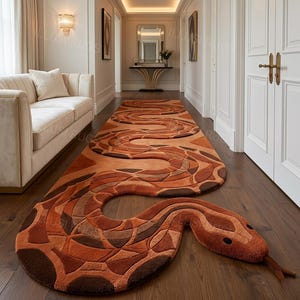 May include: A long, runner-style area rug shaped like a snake, featuring a textured surface with a repeating pattern of orange, rust, and brown geometric scales. The rug has a distinct head with dark eyes and a protruding tongue, laid on a floor.