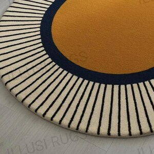 May include: A round rug with a mustard yellow center, a navy blue border, and a cream-colored outer ring with black vertical stripes. The rug has a soft, textured appearance and a modern design.