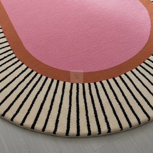 Puede incluir: Una alfombra redonda con un centro rosa, un borde color &oacute;xido y una secci&oacute;n exterior crema con rayas verticales negras. El dise&ntilde;o de la alfombra es moderno y geom&eacute;trico, adecuada para una sala de estar o dormitorio.