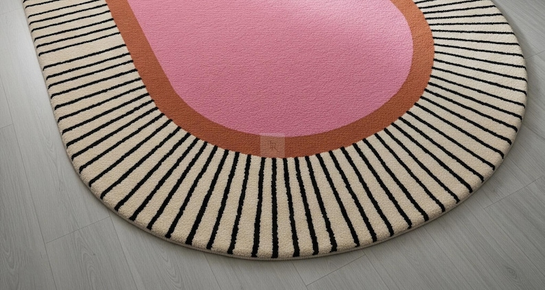 Puede incluir: Una alfombra redonda con un centro rosa, un borde color &oacute;xido y una secci&oacute;n exterior crema con rayas verticales negras. El dise&ntilde;o de la alfombra es moderno y geom&eacute;trico, adecuada para una sala de estar o dormitorio.