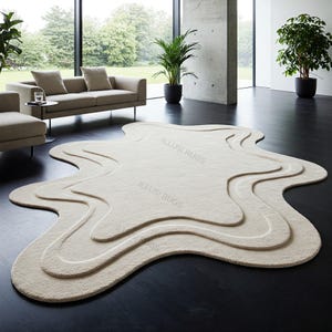 Könnte beinhalten: Ein großer, cremefarbener, flauschiger Teppich mit einem einzigartigen, abstrakten, flüssigkeitsartigen Design. Der Teppich hat drei Schichten aus erhabenen, wellenförmigen Kanten, die einen 3D-Effekt erzeugen. Der Teppich liegt auf einem dunklen Holzboden.