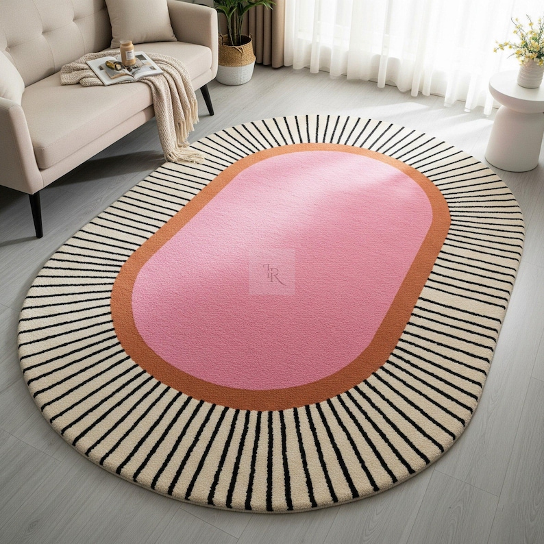Puede incluir: Alfombra ovalada con centro rosa, bordeada por una banda color &oacute;xido y un campo crema con rayas verticales negras. La alfombra est&aacute; colocada sobre un suelo claro, con un sof&aacute; y una mesa auxiliar al fondo.