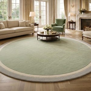 May include: A large circular area rug featuring a solid sage green center, a thin cream-colored inner border, and a wider light gray outer border. The rug is positioned on a light brown wooden floor with a herringbone pattern in a living room.