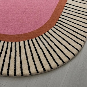 Puede incluir: Una alfombra redonda con un centro rosa, un borde curvo marr&oacute;n y un borde exterior crema con rayas verticales negras. La alfombra est&aacute; sobre un suelo de madera gris claro.