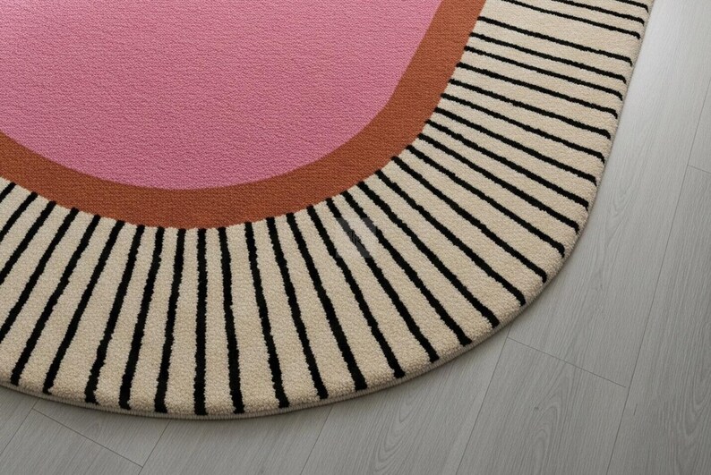Puede incluir: Una alfombra redonda con un centro rosa, un borde curvo marr&oacute;n y un borde exterior crema con rayas verticales negras. La alfombra est&aacute; sobre un suelo de madera gris claro.