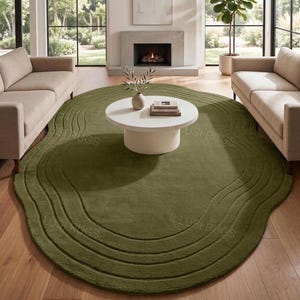 May include: An olive green, irregularly shaped area rug with a layered, wavy design. The rug is placed in a living room setting with a white coffee table, two beige sofas, and a fireplace.