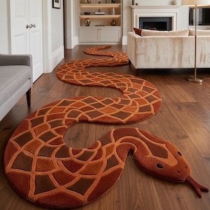 Puede incluir: Una alfombra larga y sinuosa con forma de serpiente, que presenta un patrón de diamantes repetitivo en tonos naranja quemado, óxido y marrón oscuro. La alfombra tiene una cabeza redondeada con dos ojos negros y una lengua bífida saliente.