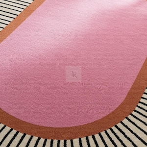 Puede incluir: Una alfombra moderna con un dise&ntilde;o &uacute;nico. La alfombra presenta una gran forma org&aacute;nica rosa en el centro, bordeada por una banda marr&oacute;n. El borde exterior tiene un patr&oacute;n de rayas blancas y negras, creando un contraste llamativo. La alfombra est&aacute; hecha de un material suave y texturizado.