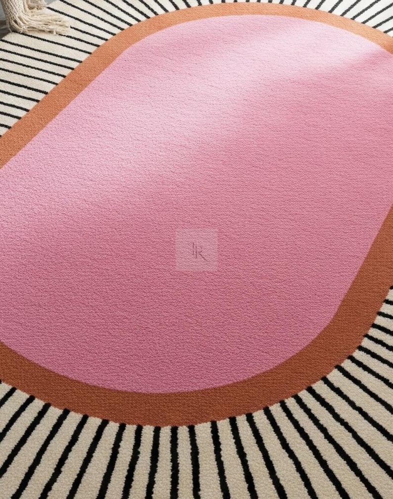 Puede incluir: Una alfombra moderna con un dise&ntilde;o &uacute;nico. La alfombra presenta una gran forma org&aacute;nica rosa en el centro, bordeada por una banda marr&oacute;n. El borde exterior tiene un patr&oacute;n de rayas blancas y negras, creando un contraste llamativo. La alfombra est&aacute; hecha de un material suave y texturizado.