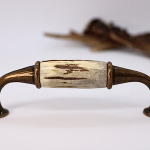 以下が含まれることがあります： 自然な角の中心部とブロンズ色の金属製金具を備えたキャビネットプル。ハンドルは、中央の角の部分とブロンズリングを備えた湾曲したデザインです。ハードウェアはヴィンテージの美学を持っています。