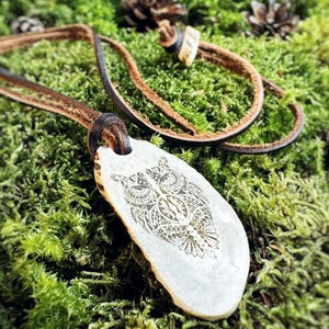 Könnte beinhalten: Ein weißer Knochenanhänger mit einem detailliert geätzten Eulenmotiv, der an einem braunen Lederband hängt. Der Anhänger liegt auf einem Bett aus grünem Moos.