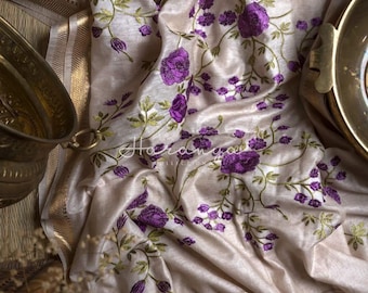 Pode incluir: Um sari de seda cor creme com uma borda dourada e bordados florais roxos. O sari está drapeado sobre uma superfície de madeira e uma tigela de latão.