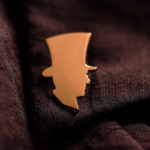 Pu&ograve; includere: Una spilla da bavero in metallo color oro a forma di silhouette di una persona che indossa un cappello a cilindro. La spilla presenta una superficie liscia e riflettente con un profilo netto e definito, che mostra un design classico in metallo.