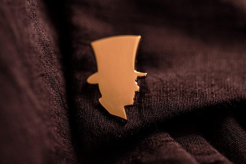 Pu&ograve; includere: Una spilla da bavero in metallo color oro a forma di silhouette di una persona che indossa un cappello a cilindro. La spilla presenta una superficie liscia e riflettente con un profilo netto e definito, che mostra un design classico in metallo.