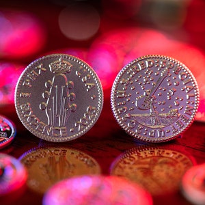 Może przedstawiać: Dwie srebrne monety z rozmytym czerwono-niebieskim tłem. Jedna moneta przedstawia gitarę z napisem "SIXPENCE" i "E.II.R.  USA  2023". Druga moneta przedstawia gitarę z napisem "SPECIAL  LIMITED  EDITION  2023  UP-1".