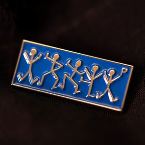 Pu&ograve; includere: Una spilla smaltata rettangolare con uno sfondo blu vivace e cinque figure stilizzate color oro in varie pose di danza. La spilla presenta un sottile bordo in metallo dorato che incornicia le figure, raffigurate in uno stile lineare.