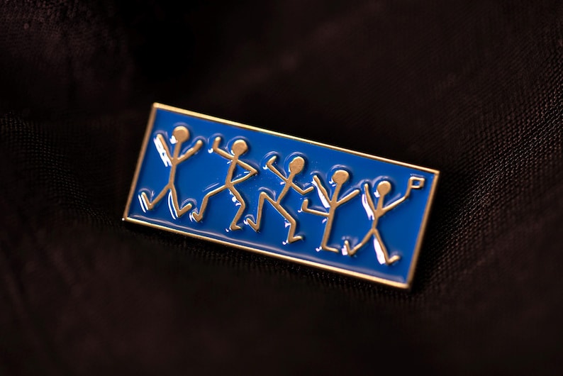 Pu&ograve; includere: Una spilla smaltata rettangolare con uno sfondo blu vivace e cinque figure stilizzate color oro in varie pose di danza. La spilla presenta un sottile bordo in metallo dorato che incornicia le figure, raffigurate in uno stile lineare.