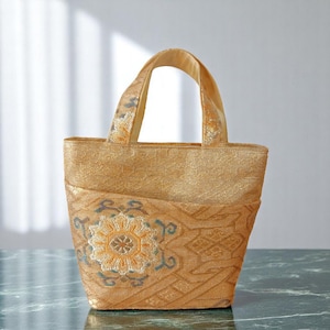 以下が含まれることがあります： 金色のトートバッグ。前面に小さなポケットが付いた長方形の形状で、花柄のデザインが施されています。花柄は黄色、青、白の色合いです。2つのハンドルがあり、テクスチャード加工された生地で作られています。