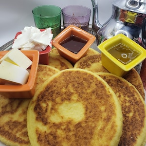 May include: A platter of golden brown flatbreads, called msemen, with a side of butter, honey, and cream cheese. The platter is set on a silver tray with colorful glasses and bowls.