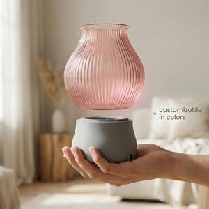 Peut inclure: Un diffuseur en verre rose avec des rainures verticales, flottant au-dessus d'une base grise. Le texte "customizable in colors" est visible. Le produit est tenu dans la main d'une personne. C'est un objet de décoration.