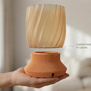 Puede incluir: Una maceta de dos piezas, de color terracota, con una base acanalada y una parte superior translúcida en espiral. Se incluye el texto "customizable in colors". La maceta se sostiene en una mano.