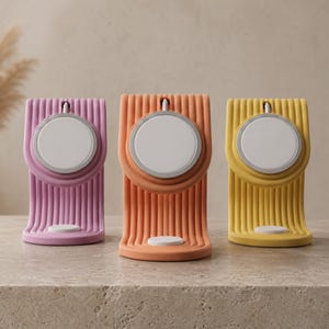 Könnte beinhalten: Drei farbenfrohe Apple Watch Ladestationen in Pink, Orange und Gelb. Jeder Ständer hat ein geripptes Design und ein kreisförmiges Ladepad. Die Ständer werden auf einer neutralen Oberfläche präsentiert.