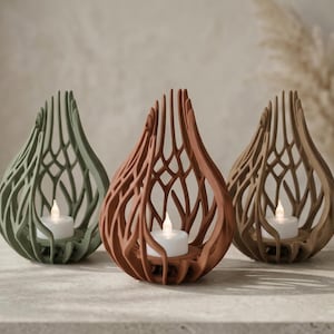 Op de afbeelding: Drie decoratieve kandelaars in groen, terracotta en bruin. Elke houder heeft een druppelvorm met een open, tralieachtig ontwerp. In elke houder zit een witte LED-kaars.
