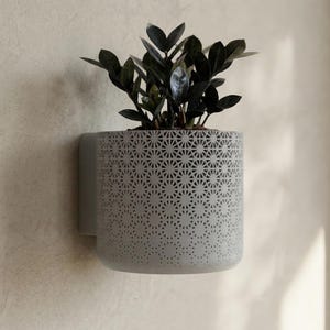 Peut inclure: Un pot mural gris avec un motif géométrique, contenant une plante vert foncé. Le pot est fixé à un mur de couleur claire. Le pot cylindrique présente des découpes en forme d'étoile.