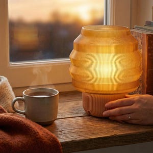 Può includere: Una lampada ambrata e luminosa con un design a coste su un davanzale di legno. Una tazza di tè, una coperta color crema e libri d'epoca sono nelle vicinanze. La scena crea un'atmosfera accogliente. La lampada è un elemento di illuminazione decorativo.