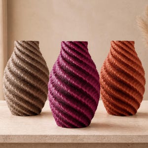 Könnte beinhalten: Drei Vasen mit einer ausgeprägten spiralförmigen Rippenstruktur. Die linke Vase ist in einem gedeckten Taupe gehalten, die mittlere Vase in einem tiefen Magenta und die rechte Vase in einem warmen Terrakotta-Orange. Jedes Gefäß hat einen Hals.