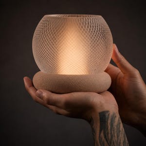 May include: A spherical lamp with a woven design, emitting a warm glow. The lamp sits on a circular base and is held in open hands. The background is dark.