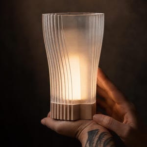May include: A frosted glass lamp with vertical ridged detailing and a bronze-coloured base. The lamp is lit from within, casting a warm glow. The lamp is held in a hand against a dark background.