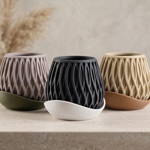 May include: Three decorative planters in a row. The planters are in shades of mauve, black, and beige, each with a unique base color. The planters have a woven design and are suitable for indoor plants.