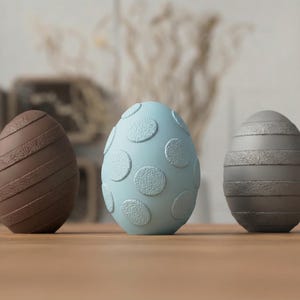 Può includere: Tre uova di Pasqua decorative. Il primo è marrone con strisce diagonali strutturate. L'uovo centrale è azzurro con grandi cerchi strutturati. L'ultimo uovo è grigio con strisce orizzontali strutturate. Tutte le uova sono su una superficie di legno.