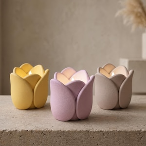 Può includere: Tre portacandele a forma di tulipano in giallo, lavanda e beige. Ogni portacandele ha una candela accesa all'interno, che emana un bagliore caldo. I portacandele sono realizzati in materiale opaco e poggiano su una superficie di pietra. Arredamento per la casa.