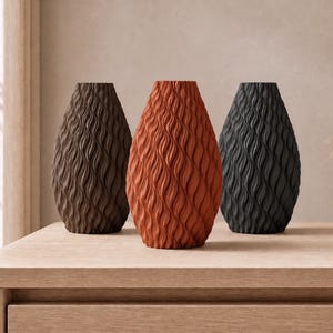 Pode incluir: Três vasos em forma de gota com uma textura ondulada vertical. O vaso à esquerda é castanho escuro, o vaso central é laranja terracota e o vaso à direita é preto mate. Cada vaso apresenta um padrão repetitivo consistente.