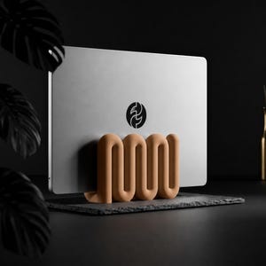 Op de afbeelding: Een zilveren laptop met een zwart cirkelvormig logo met twee gestileerde witte vormen wordt rechtop gehouden door een matte, beige, golvende standaard. De standaard heeft een doorlopend, herhalend S-vormig ontwerp dat het apparaat ondersteunt.